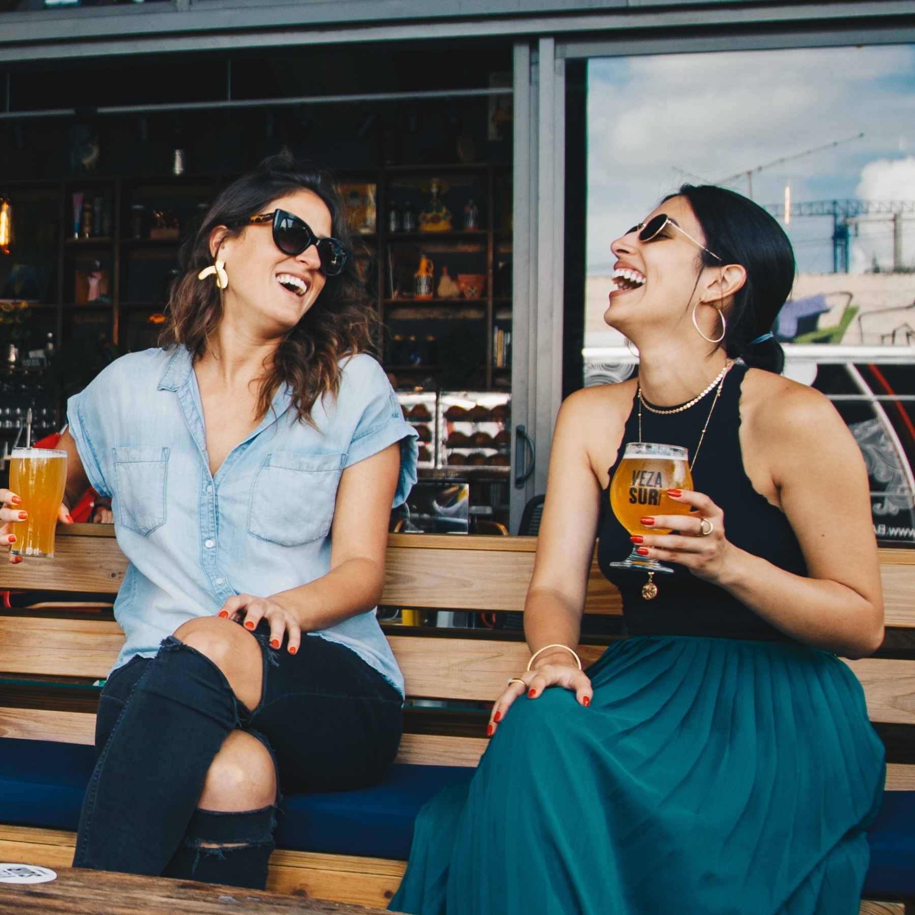 Friends seated on bench at brewery with beer glasses laughing.